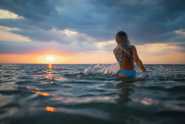 Sarı saçlı, mavi mayolu bir kız gün batımı arka planında bir nehir kenarında otururken kenarlara su sıçratıyor. — Stok fotoğraf