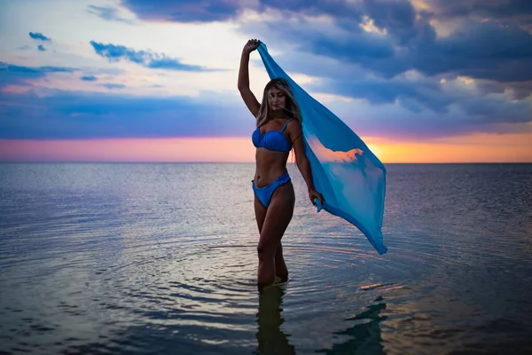 A girl in a blue swimsuit and a bright pareo posing against the background of a sunset in estuary with transparent water — Stock Photo, Image