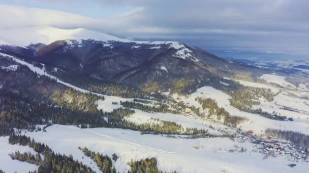 Mała, pokryta śniegiem wioska w ogromnej górskiej dolinie z lasami świerkowymi na tle zachmurzonego błękitnego nieba — Wideo stockowe