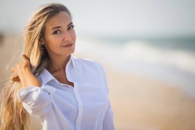 Uzun saçlı, mavi mayolu ve gömlekli bir kız deniz kıyısında saçlarını tarıyor.