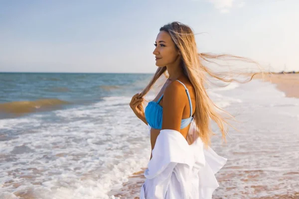 Стройная девушка в мягком голубом купальнике и рубашке прогуливается по песчаному пляжу у синего моря — стоковое фото