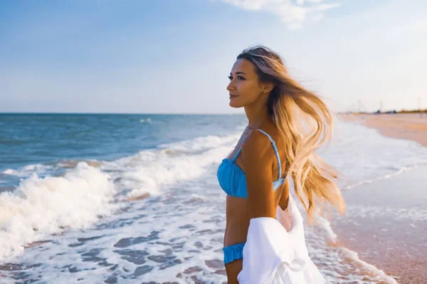 Uma menina delgada em um terno de banho azul suave e camisa caminha ao longo da praia de areia perto do mar azul — Fotografia de Stock