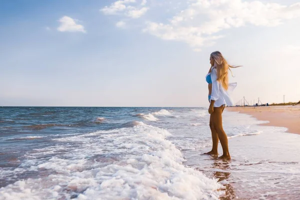 Una chica delgada en un traje de baño azul suave y camisa camina a lo largo de la playa de arena cerca del mar azul —  Fotos de Stock