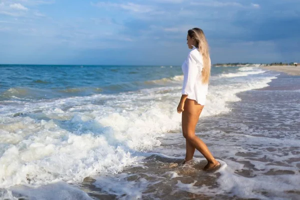 Uma menina delgada em um terno de banho azul suave e camisa caminha ao longo da praia de areia perto do mar azul — Fotografia de Stock