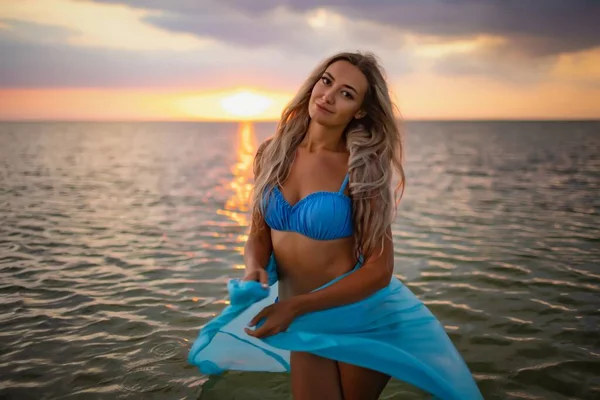 Una chica en traje de baño azul y un brillante pareo posando sobre el fondo de una puesta de sol en el estuario con agua transparente —  Fotos de Stock