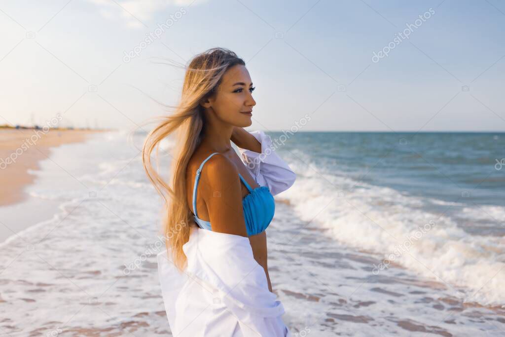 A slender girl in a gentle blue swimsuit and shirt walks along the sandy beach near the blue sea