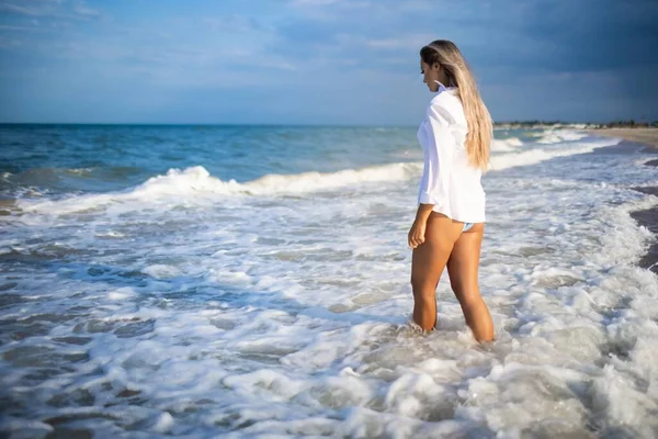 Una chica delgada en un traje de baño azul suave y camisa camina a lo largo de la playa de arena cerca del mar azul —  Fotos de Stock