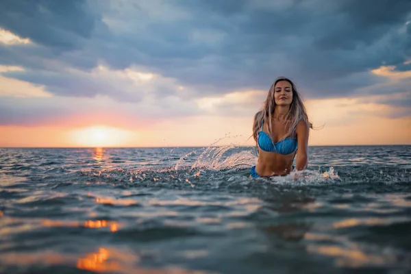 A girl with blond hair in a blue swimsuit splashes to the sides while sitting in an estuary on a sunset background — Stock Photo, Image