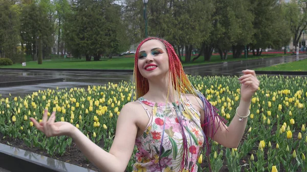 漂亮的女孩与彩虹辫子和表达华丽的妆容 她坐在一个绿树成荫的公园里 享受着春天的温暖天气 — 图库照片