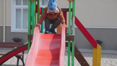 Sıcak giysili, mavi şapkalı, neşeli, hareketli bir çocuk bir anaokulunda çocuk parkında oynuyor ve sonbahar bulutlu bir havada çok renkli bir slaytta kayıyor. HD yavaş çekim videosu