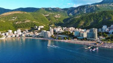 Budva ve Becici tatil beldeleri, sakin Adriyatik Denizi yakınlarında lüks oteller ve rahat plajlarla Karadağ Dağları ve mavi gökyüzüne karşı. UHD Video gerçek zamanlı 4K