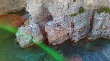 Saf gri kayalıklar, denizin üzerinde tehlikeli keskin yamaçlar, mavi renkli sakin dalgalar, resiflerle kaplı deniz yatakları, büyük kaldırım taşları ve kum. UHD Video gerçek zamanlı 4K