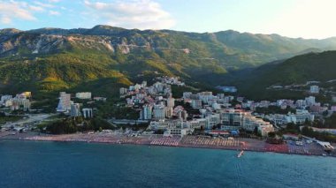 Geniş plaj, güneş şemsiyeleri, plastik beyaz güneşlikler ve Adriyatik Denizi yakınlarındaki Becici tatil beldesinde dinlenen insanlar akşam güneşli gökyüzüne karşı. UHD Video gerçek zamanlı 4K