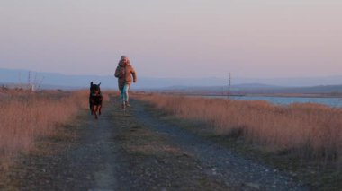 Ceketli enerjik neşeli genç kız mavi deniz kenarındaki kumlu sahilde sevgili sadık arkadaşı Rottweiler cinsinin büyük mutlu köpeği 4K UHD yavaş çekim videosu