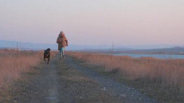 Ceketli enerjik neşeli genç kız mavi deniz kenarındaki kumlu sahilde sevgili sadık arkadaşı Rottweiler cinsinin büyük mutlu köpeği 4K UHD yavaş çekim videosu