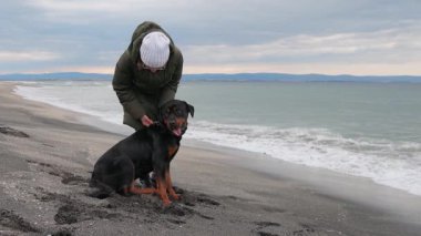 Sıcak ceketli bilinmeyen bir kız mavi fırtınalı denizin yanındaki kumlu sahilde duruyor ve sadık arkadaşının kulağının arkasını tırmalıyor. Rottweiler cinsinin büyük, eğitimli köpeği. 4K UHD yavaş çekim videosu
