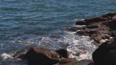 Bulgaristan 'ın parlak güneşli günbatımı ışığı altında, Karadeniz' in yansıtıcı ilkbaharı yakınlarında, koyu nemli deniz kumları ve büyük ıslak kayalık kayalıkları olan vahşi bir taş ve kumlu bir sahil. 4K UHD yavaş çekim videosu