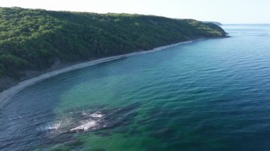 A wild empty sandy beach between a rocky sharp large ledge, covered with green fresh new vegetation, and a deep calm cool Black Sea under a clear blue sky. UHD 4K video realtime