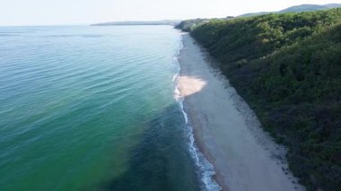 A wild empty sandy beach between a rocky sharp large ledge, covered with green fresh new vegetation, and a deep calm cool Black Sea under a clear blue sky. UHD 4K video realtime