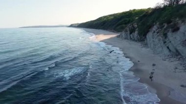 Rocky high sharp ledge with new fresh green vegetation over wild empty sandy beach in the shadow of a rock, near the deep calm undulating Black Sea under a clear blue bright sky. UHD 4K video realtime