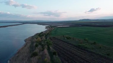 Calm bluish water surface of Black Sea washes wild coast and green farmer fields in valley of Bulgaria, against background of cloudy color vast sky with bright sunset sun. UHD 4K video realtime
