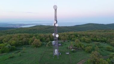Birds eye view of hills covered with green spring vegetation with tall professional cell towers, against the backdrop of farmhouse blooming planted fields and a clear blue sky. UHD 4K video realtime