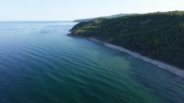 A wild empty sandy beach between a rocky sharp large ledge, covered with green fresh new vegetation, and a deep calm cool Black Sea under a clear blue sky. UHD 4K video realtime