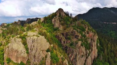 High rocky wild mountain range of Rhodope Mountains covered with green vegetation, against backdrop of mountain valley covered with dense dark spruce forests and blue cloudy sky. UHD 4K video realtime