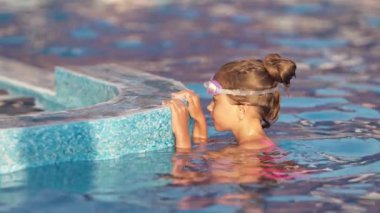 Yüzmek için şeffaf siyah gözlüklü, parlak pembe bir mayo giyen küçük bir kız gözlüklerini düzeltiyor ve berrak bir su şemsiyesiyle birlikte derin mavi bir havuza dalıyor. 4K UHD yavaş çekim videosu