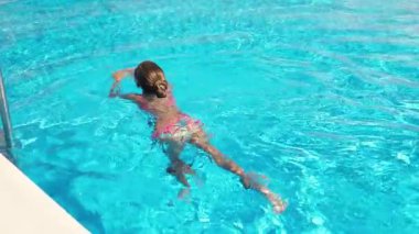 Güzel, komik, ince, parlak pembe bir mayo giyen mor gözlüklü bir kız ılık bir yaz gününde şeffaf suyla dolu derin bir havuzda süzülüyor.