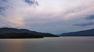 Small blue reflecting lake and clear cool water with stone narrow wild shore in coniferous spruce forest with tall fir trees, against pale blue daytime cloudy sky. UHD 4K video realtime