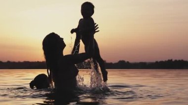 Mutlu anne, parlak altın yaz güneşli günbatımına karşı şeffaf, berrak, berrak gölde renkli kırmızı kolluklarıyla gülen küçük çıplak bebekle oynuyor. HD video yavaş çekim