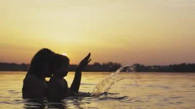 Mutlu bir anne ve çıplak gülen bir bebeği var. Kucağında kızıyla oynuyor ve açık mavi gölde parlak güneşli yaz günbatımının arka planına doğru sıçrıyor. HD video yavaş çekim