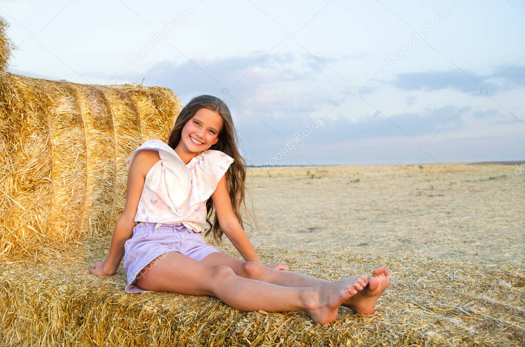 Adorable happy smiling ittle girl child sitting on a hay rolls in a wheat field at sunset