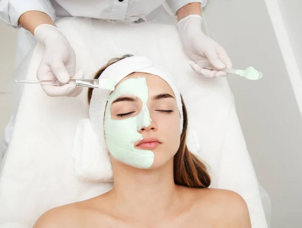 Beautiful young woman with facial mask in beauty salon. Girl getting beauty treatment facial care by beautician