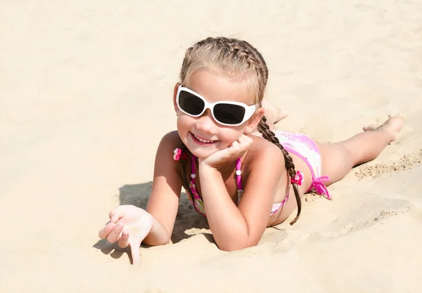 Happy cute little girl in sunglasses lying on the sand — Stock Photo, Image