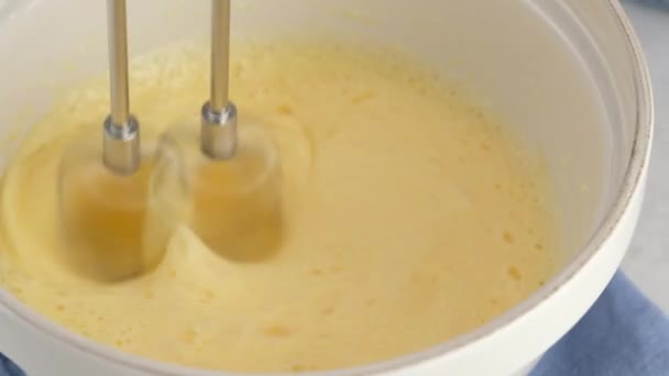 Primer plano mezclando claras de huevo, yemas y azúcar en un tazón de cerámica con una batidora manual sobre una mesa blanca. Cocinar un bizcocho. — Vídeos de Stock