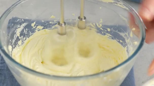 白いテーブルの上に電動ミキサーでクリームを作るためのガラスボウルに女性の手ホイップバターのクローズアップ.シャルロットクリームでスポンジケーキを作る. — ストック動画