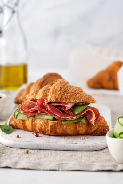 Čerstvý croissant nebo sendvič s okurkou, šunkou, jamonem, prosciuttem, na dřevěném podkladu. Ranní snídaně. Zdravé jídlo. — Stock fotografie