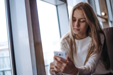 Üzgün sarışın kadın kanepede oturuyor ve akıllı telefon kullanıyor.
