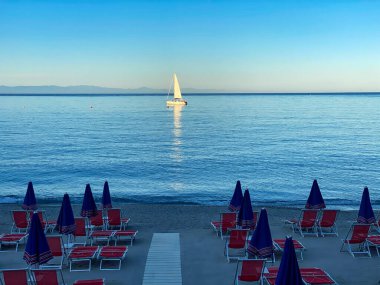 Sahilin önündeki körfezde demirlemiş bir yelkenli üzerinde şemsiyeler ve straio, Noli, Liguria - İtalya