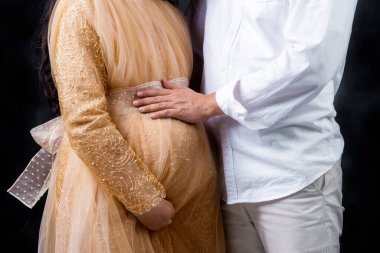 Hamile çift, elleri siyah arka planda, göbeğine dokunuyor.