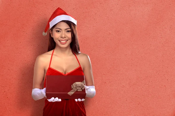 身穿圣诞老人服装的亚洲女人拿着红墙背景的礼品盒 圣诞快乐 — 图库照片