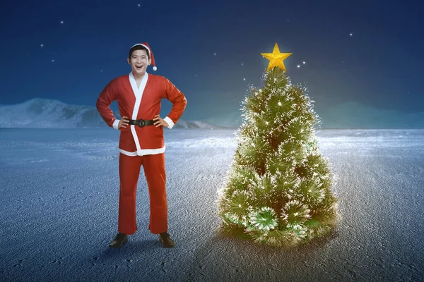サンタの衣装を着たアジア人男性が夜の風景を背景にクリスマスツリーに立っています メリークリスマス — ストック写真