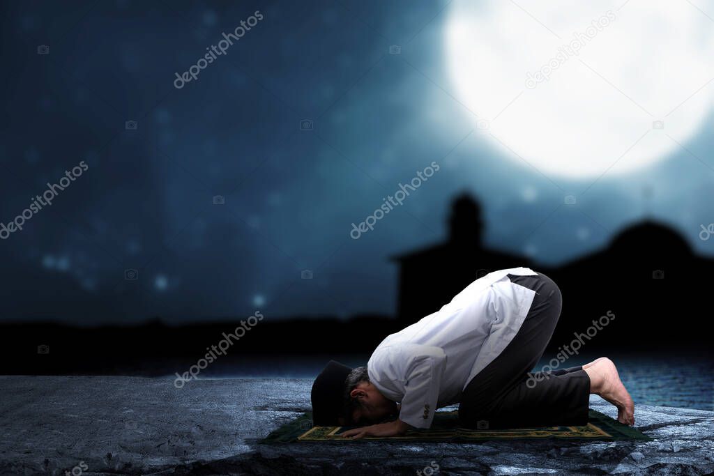 Asian Muslim man in praying position (salat) on prayer rug with the night scene background