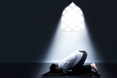 Dua eden Asyalı Müslüman bir adam (namaz kılma pozisyonu) ışık arka planda, odanın içindeki halının üzerinde.