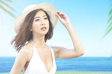 Bikinili ve şapkalı Asyalı kadın okyanus manzaralı sahada dinleniyor.