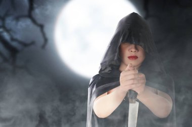 Pelerinli Asyalı cadı elinde bıçakla sahne arkasında duruyor.