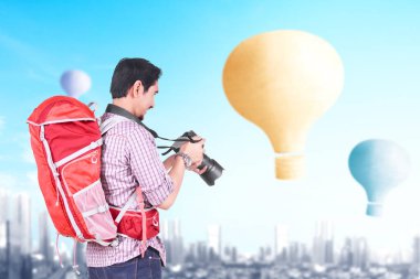 Sırt çantalı ve kameralı Asyalı adam şehir planıyla uçan renkli hava balonuna bakıyor