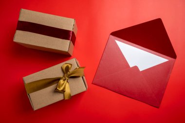 Kahverengi hediye kutusunun üst görüntüsü altın ve kırmızı kurdeleyle bağlanmış ve arka planda kırmızı zarf var. Model ya da kopyalama alanı. Yıldönümleri, kutlamalar, hediyeler veya sürprizler için dekorasyon kavramı